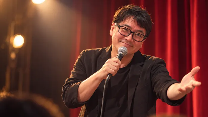 赤い緞帳の舞台でマイクを握り、身ぶりを交えて笑いながら話す浦井崇