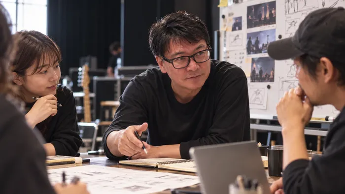 絵コンテの貼られた制作現場で、机を囲んでスタッフと話す浦井崇